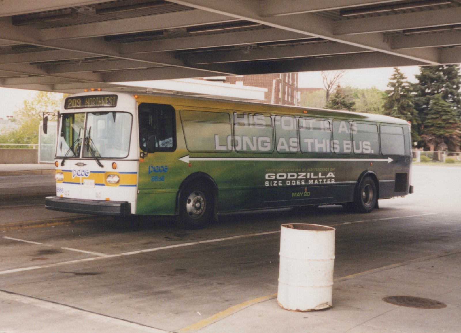 A white and green Pace bus numbered 8866, parked under a concrete overpass, features a large movie ad for 'Godzilla' on its side and shows route 209 Northwest on its front sign.