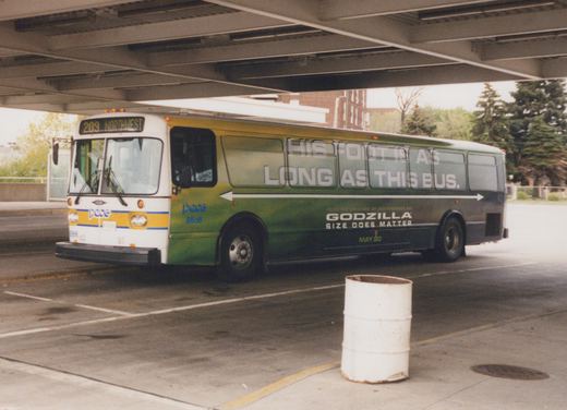 A white and green Pace bus numbered 8866, parked under a concrete overpass, features a large movie ad for 'Godzilla' on its side and shows route 209 Northwest on its front sign.