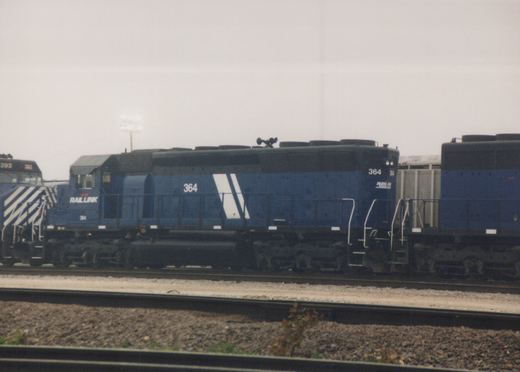 Side view of a blue Rail Link locomotive number 364, with another locomotive numbered 393 behind it, both sitting on railroad tracks in a yard on an overcast day.