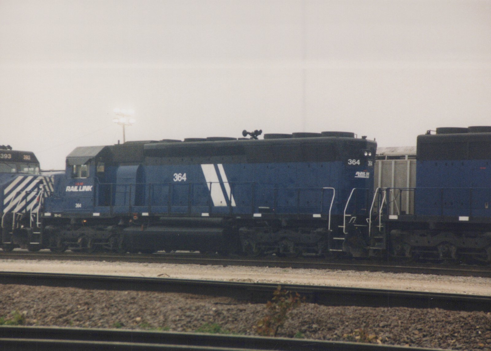 Side view of a blue Rail Link locomotive number 364, with another locomotive numbered 393 behind it, both sitting on railroad tracks in a yard on an overcast day.