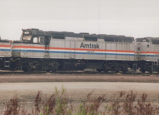 A side view of two Amtrak locomotives, numbers 302 and 345, painted in silver with red and blue stripes, sitting on railroad tracks in a yard.