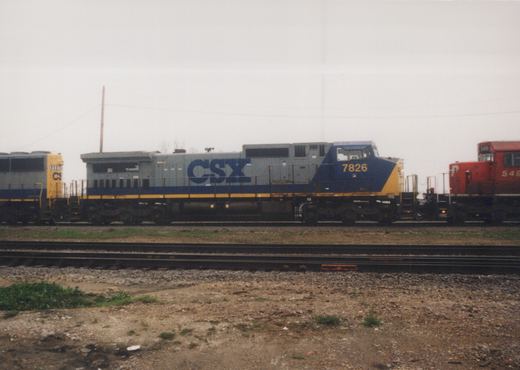 Three diesel locomotives are parked on parallel tracks in a gravel yard under an overcast sky. The central locomotive is gray and blue with CSX lettering and the number 7826. To its left is another CSX unit, and to its right is a red Penn Central locomotive numbered 549.