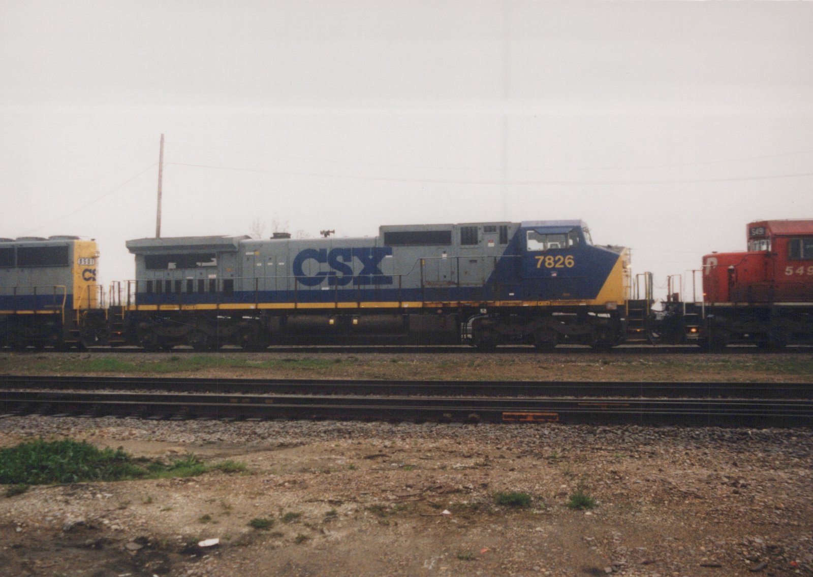 Three diesel locomotives are parked on parallel tracks in a gravel yard under an overcast sky. The central locomotive is gray and blue with CSX lettering and the number 7826. To its left is another CSX unit, and to its right is a red Penn Central locomotive numbered 549.
