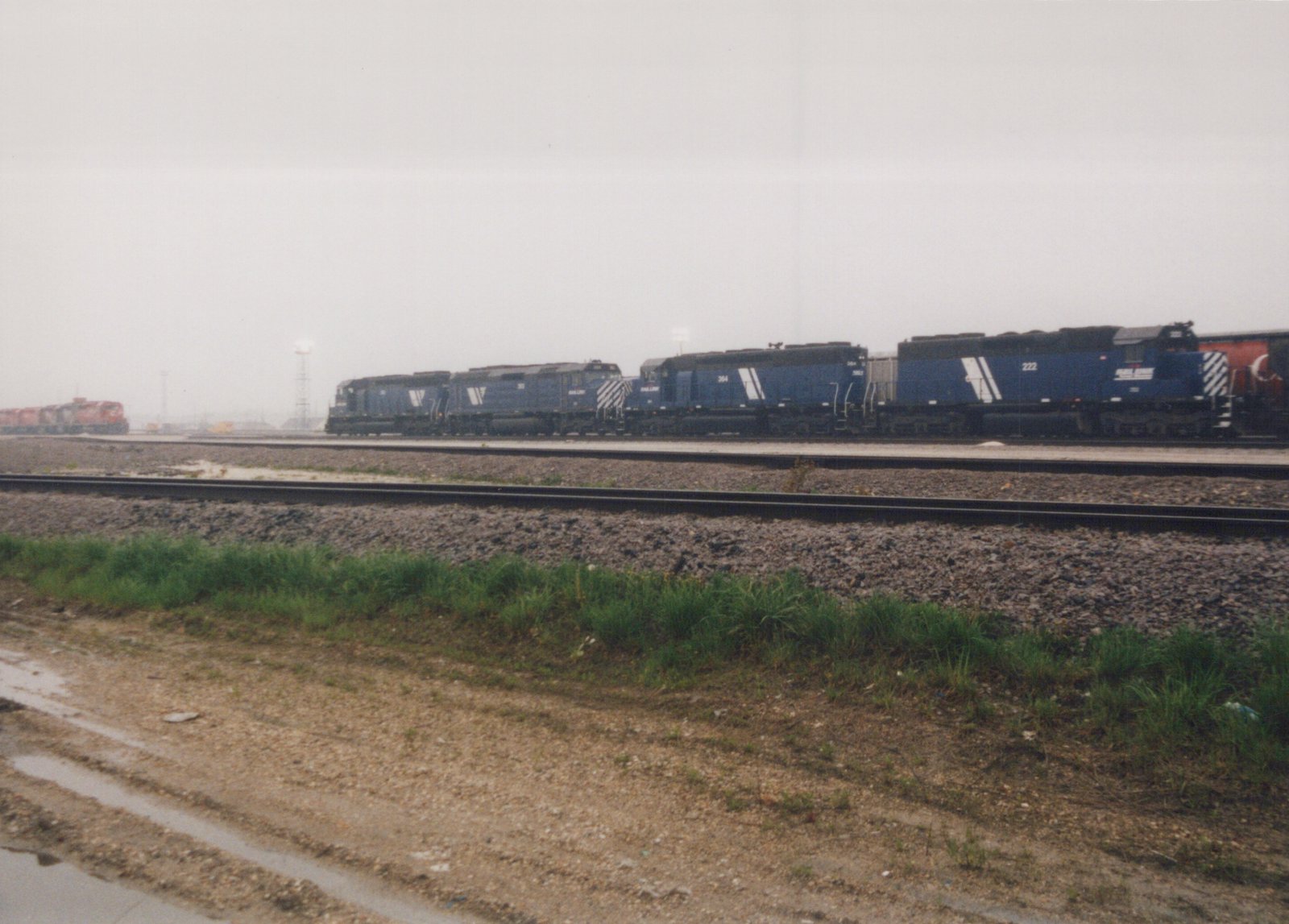 A long line of dark blue Wabash GP9 diesel locomotives, including numbers 222 and 364, are parked on multiple tracks in a gravel rail yard under a gray, overcast sky.