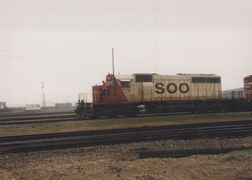 A SOO Line diesel locomotive numbered 775 stands in a rail yard, with a person visible on its side, surrounded by tracks and freight cars under a cloudy sky.