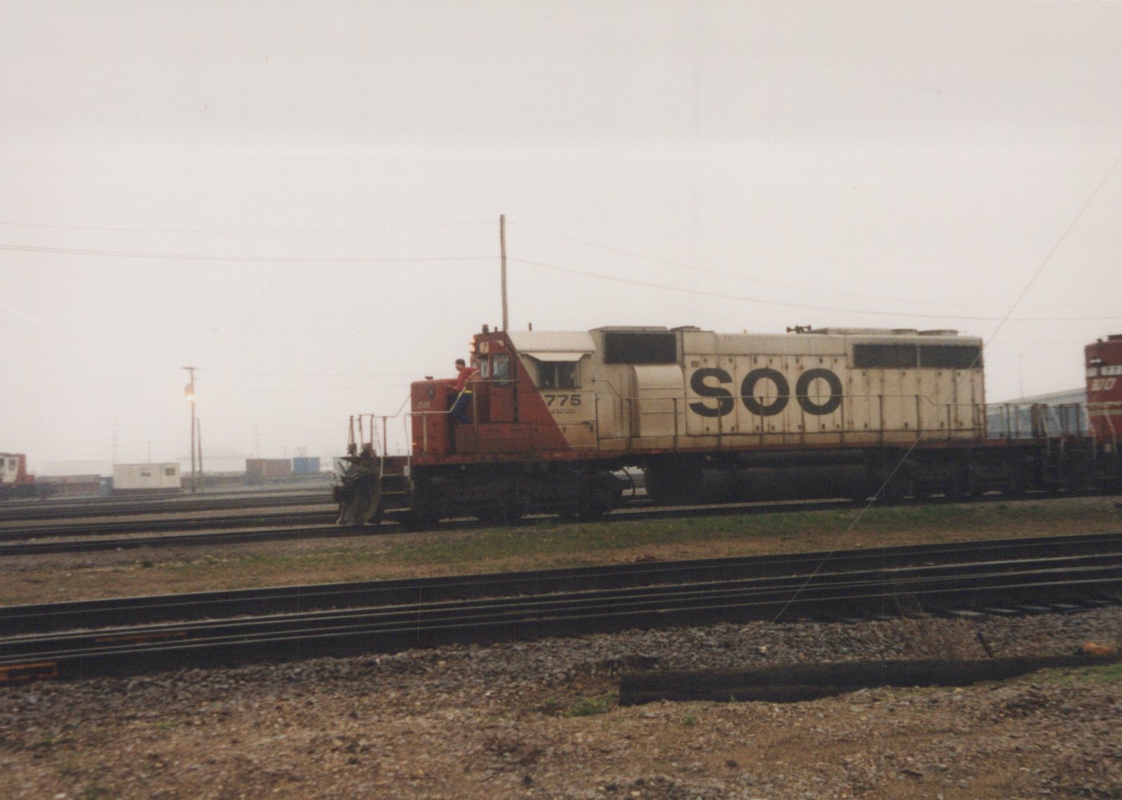 A SOO Line diesel locomotive numbered 775 stands in a rail yard, with a person visible on its side, surrounded by tracks and freight cars under a cloudy sky.