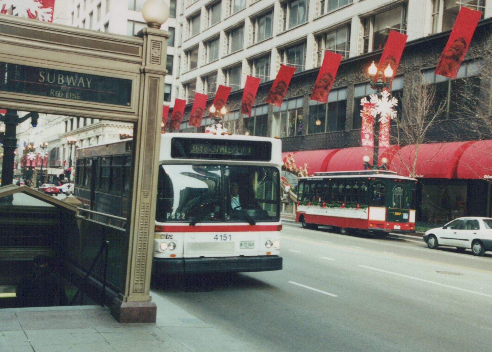 A white and red CTA bus and a green and red vintage streetcar are driving on a city street lined with holiday banners, next to a subway entrance.