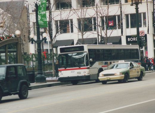 A white CTA bus with red and blue stripes, numbered 4321 and marked for route 67, is driving on a city street lined with holiday decorations and a Borders bookstore sign.