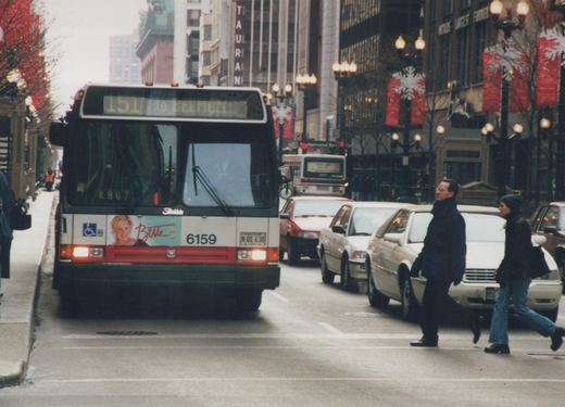 Front view of a Chicago Transit Authority bus on a snowy city street, with pedestrians crossing and holiday banners on the lampposts.