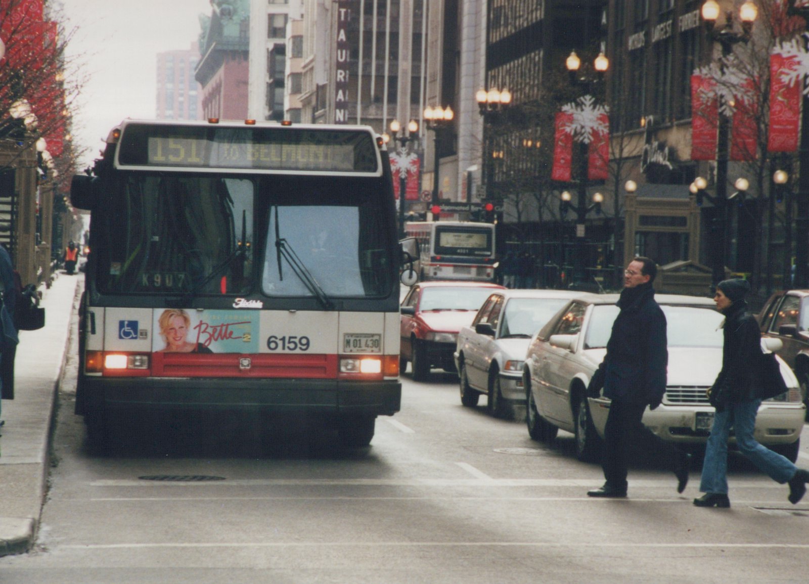 Front view of a Chicago Transit Authority bus on a snowy city street, with pedestrians crossing and holiday banners on the lampposts.