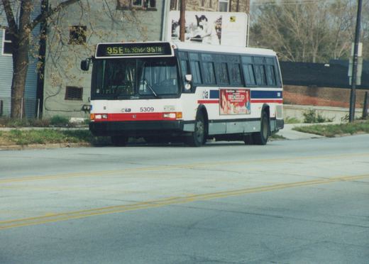A white, red, and blue CTA bus with the number 5309 is driving on a street, displaying a destination sign for route 95E to 92nd/95th. A billboard and trees are visible in the background.