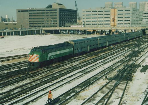 A green and white Metra locomotive numbered 9920 pulls silver commuter cars across multiple tracks in a snowy urban rail yard with modern buildings in the background.
