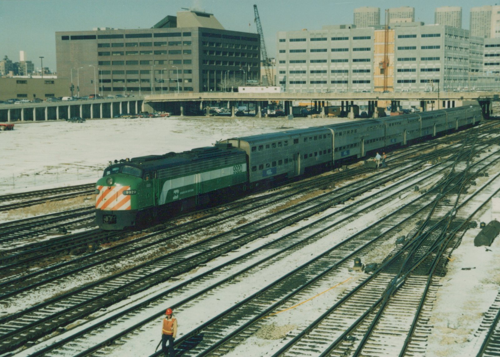 A green and white Metra locomotive numbered 9920 pulls silver commuter cars across multiple tracks in a snowy urban rail yard with modern buildings in the background.