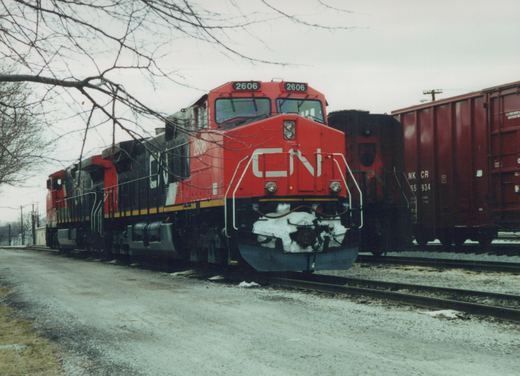 A red and black Canadian National locomotive, number 2606, is parked on a railroad track next to a brown freight car in a snowy yard with bare trees in the background.