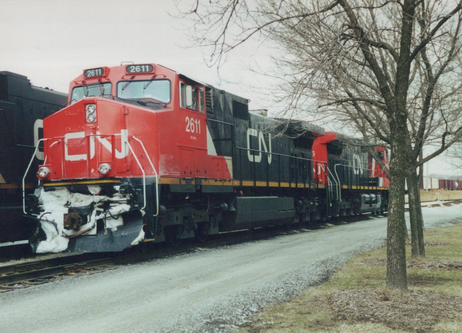A red and black Canadian National diesel locomotive, number 2611, is shown on a railroad track with snow on the ground and bare trees in the background, indicating a winter scene.