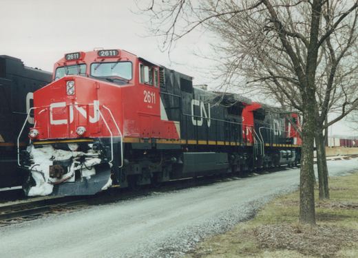 A red and black Canadian National diesel locomotive, number 2611, is shown on a railroad track with snow on the ground and bare trees in the background, indicating a winter scene.