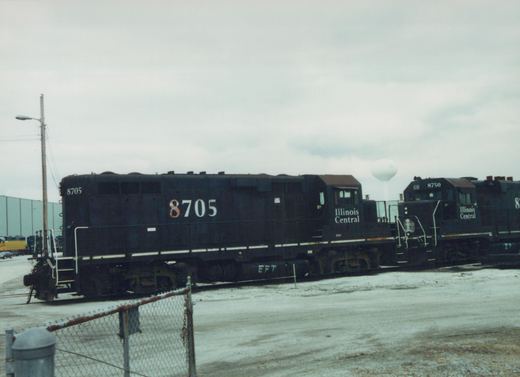 A line of two black Illinois Central diesel locomotives, numbered 8705 and 8750, are parked in a snowy yard next to a chain-link fence and a water tower.