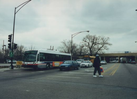 A white and blue CTA bus numbered 6468 on route 87 to 57th Street is stopped at a street intersection under an overcast sky, with a pedestrian crossing the street and other vehicles nearby.