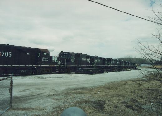 A row of dark-colored Illinois Central locomotives with numbers like 8705 and 8750 are parked side-by-side on a gravel yard under an overcast sky, with bare trees and a water tower in the background.