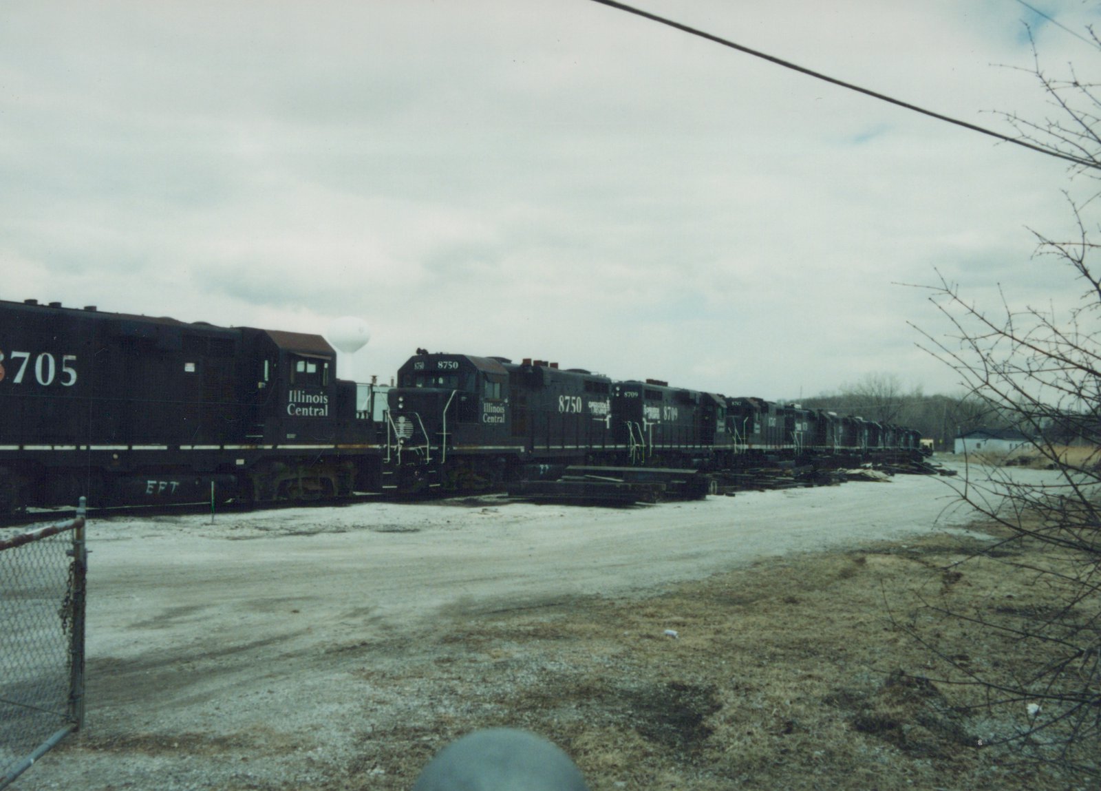 A row of dark-colored Illinois Central locomotives with numbers like 8705 and 8750 are parked side-by-side on a gravel yard under an overcast sky, with bare trees and a water tower in the background.