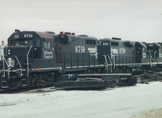 A line of three dark gray Illinois Central diesel locomotives, numbered 8750, 8709, and 8747, are parked on tracks in a snowy yard.