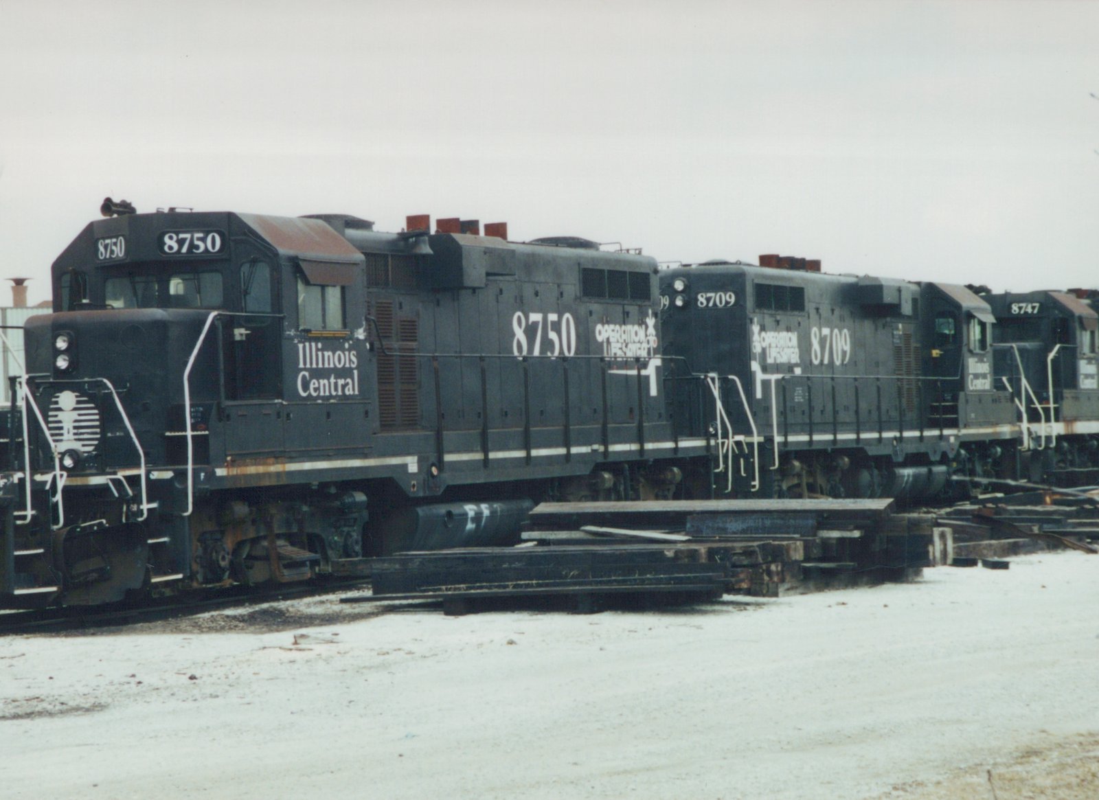 A line of three dark gray Illinois Central diesel locomotives, numbered 8750, 8709, and 8747, are parked on tracks in a snowy yard.