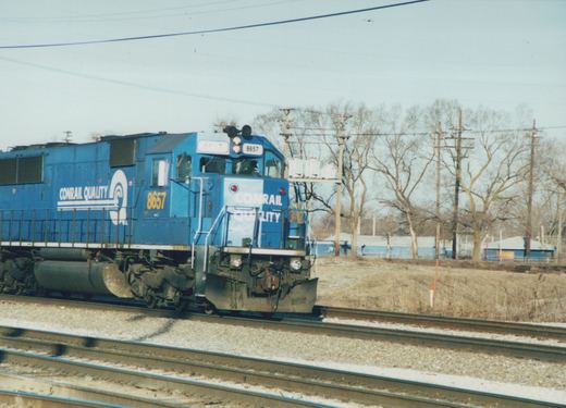 A blue Conrail diesel locomotive numbered 8657 is moving along tracks with bare trees in the background under a clear sky.