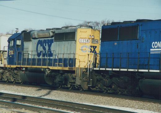 A blue and white CSX locomotive numbered 8114 on railroad tracks, with a second blue locomotive partially visible behind it, under a clear sky in winter.
