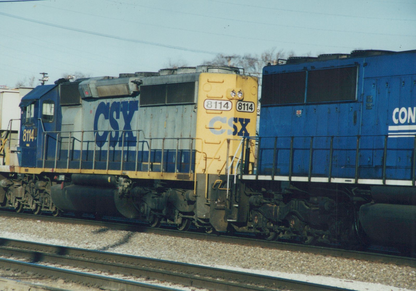 A blue and white CSX locomotive numbered 8114 on railroad tracks, with a second blue locomotive partially visible behind it, under a clear sky in winter.
