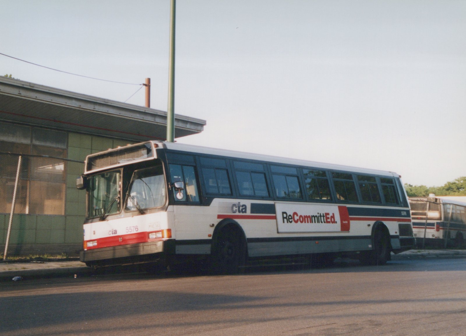 A white and blue CTA bus with the number 5576 on its front and side is parked on a paved lot next to a building, with another similar bus visible in the background.