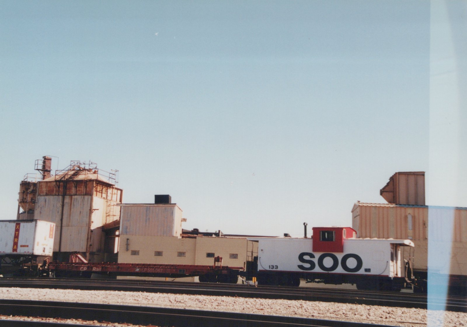 A white and red Soo Line caboose numbered 133 on railroad tracks, with industrial buildings and a CP Rail flatcar in the background under a clear sky.