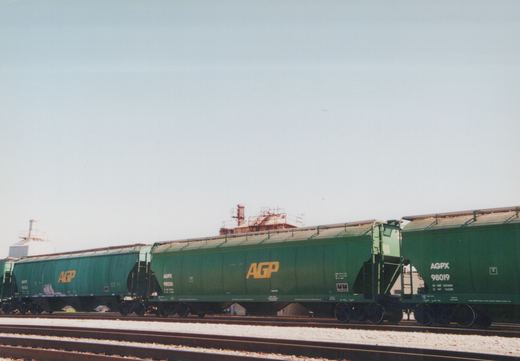A side view of three green AGPX covered hopper railcars on parallel tracks, with industrial buildings in the background under a clear sky.