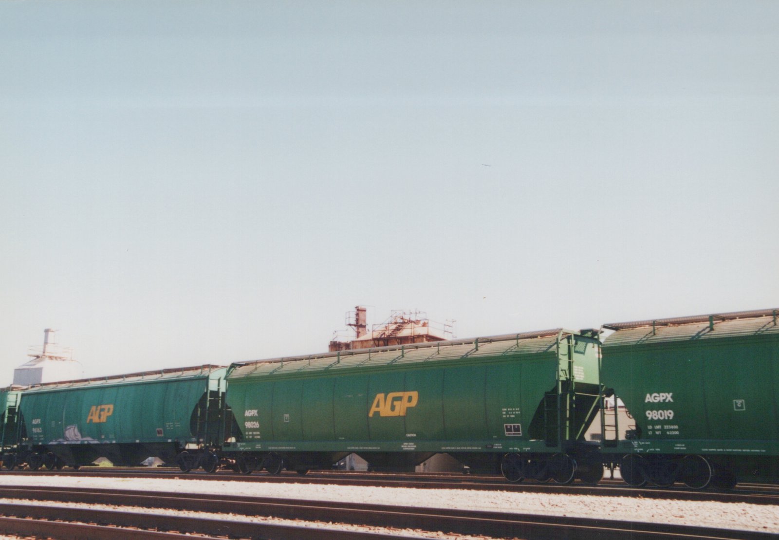 A side view of three green AGPX covered hopper railcars on parallel tracks, with industrial buildings in the background under a clear sky.