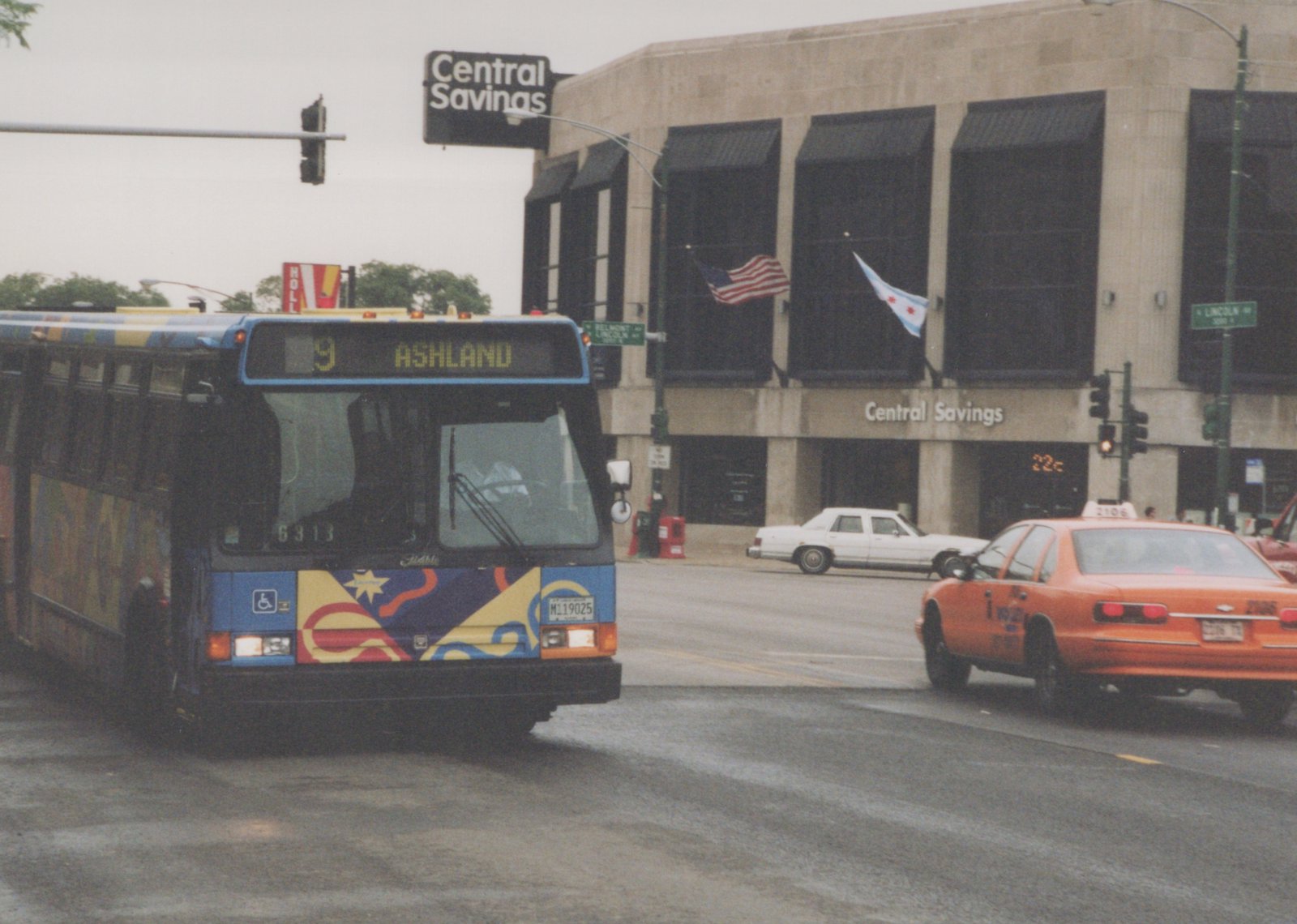 A blue and colorfully decorated CTA bus is turning at an intersection in Chicago, with a taxi and a building labeled 'Central Savings' in the background.