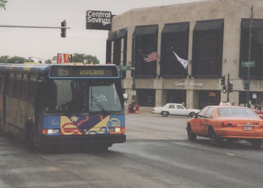 A blue and colorfully decorated CTA bus is turning at an intersection in Chicago, with a taxi and a building labeled 'Central Savings' in the background.