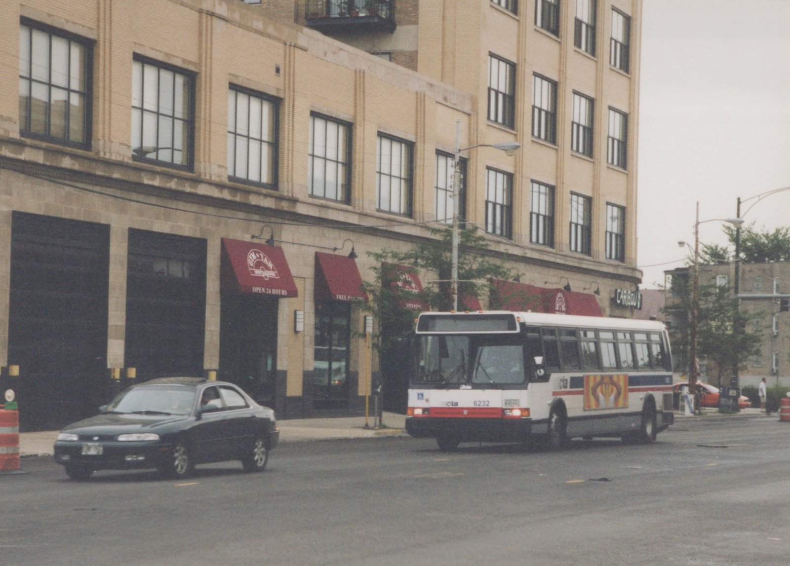 A white and red CTA bus numbered 6232 is turning a corner on a city street, with a brick building and a dark sedan in the background.