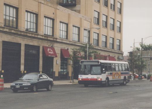 A white and red CTA bus numbered 6232 is turning a corner on a city street, with a brick building and a dark sedan in the background.