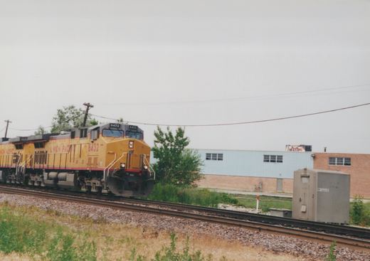 A yellow Union Pacific diesel locomotive with the number 6432 on the front is moving along a track next to a building with trees and power lines in the background.
