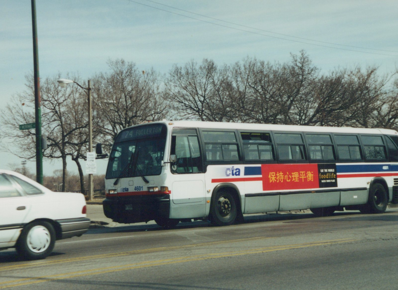 A white Chicago Transit Authority bus with red and blue stripes, fleet number 4601, displays a destination of '74 Fullerton' on its front sign, driving down a street lined with bare trees under an overcast sky.