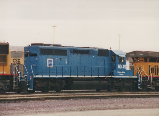 Side view of a blue Electro-Motive Leasing diesel locomotive numbered 6049 in a rail yard, with a yellow CSX locomotive behind it.