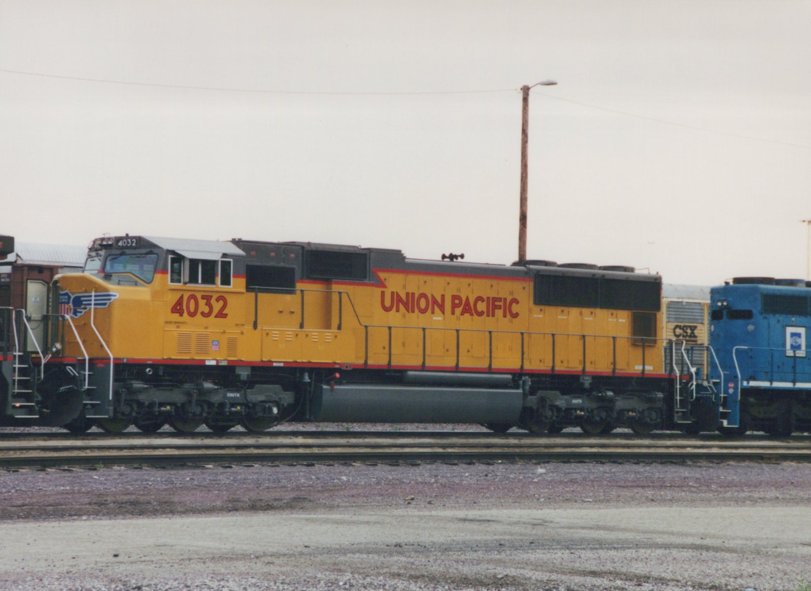 A yellow and red Union Pacific diesel locomotive, number 4032, sits on the tracks in a rail yard with other trains and a blue locomotive visible in the background.
