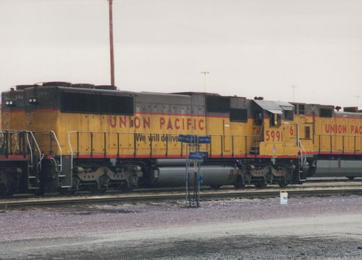 A yellow and gray Union Pacific diesel locomotive, number 5991, is parked on tracks in a yard, with its side showing the company's logo and the slogan 'We will deliver'.