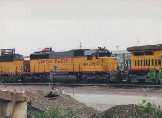 Two Union Pacific diesel locomotives, one an SD40-2 and the other a GP38-2, are parked in a gravel yard under a gray sky, with billboards visible in the background.