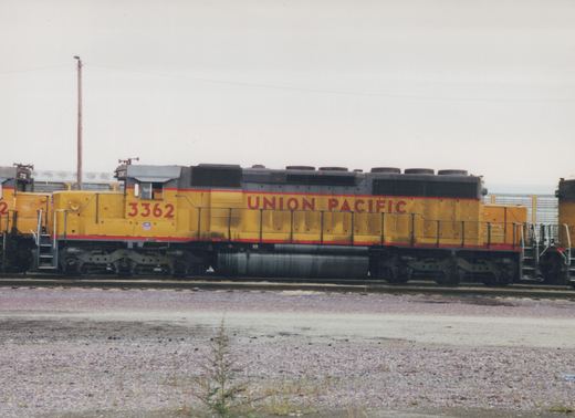 A side view of a Union Pacific diesel locomotive, number 3362, painted yellow with a gray roof and red stripe, parked on a railroad track in a gravel yard under an overcast sky.
