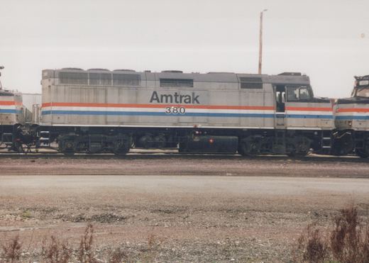 A gray Amtrak locomotive with red, white, and blue stripes, numbered 380, stands on a track in a gravel yard with a second locomotive visible on the right.