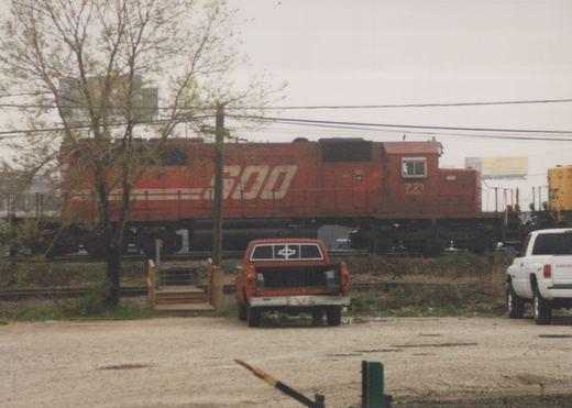 A red SOO Line locomotive numbered 771 sits on railroad tracks near a dirt road with two pickup trucks, one red and one white, parked nearby on an overcast day.