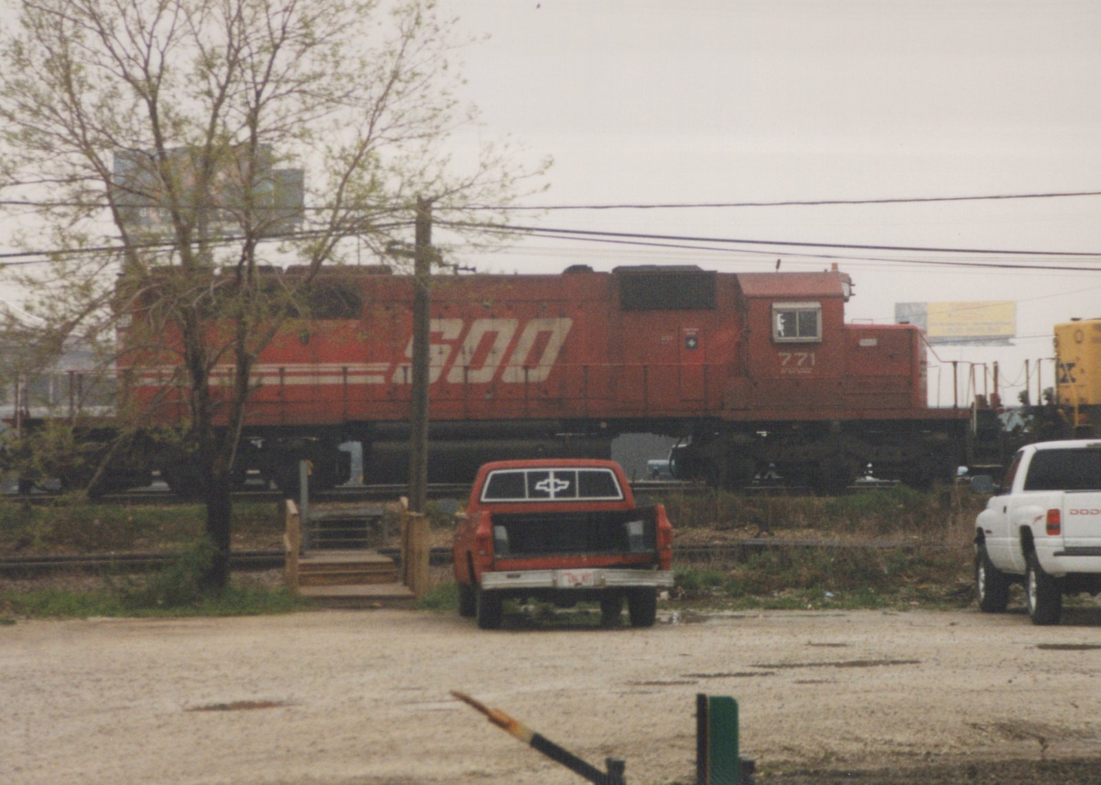 A red SOO Line locomotive numbered 771 sits on railroad tracks near a dirt road with two pickup trucks, one red and one white, parked nearby on an overcast day.