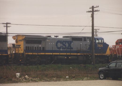 A CSX diesel locomotive numbered 7826 is parked in a freight yard next to a red Canadian Pacific locomotive numbered 50, with a black SUV and utility poles nearby under a cloudy sky.