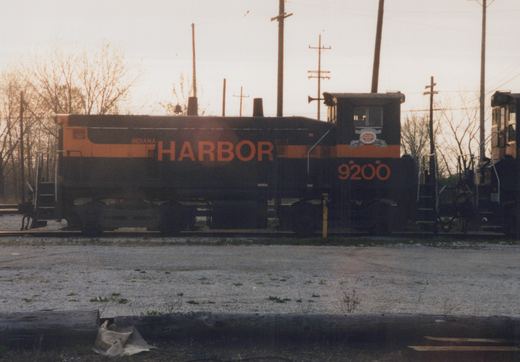 Side view of a black and orange Indiana Harbor Belt diesel locomotive, number 9200, parked on tracks in a gravel yard with bare trees and utility poles in the background.