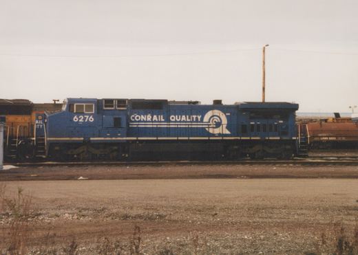 Side view of a blue Conrail locomotive number 6276, parked in a gravel rail yard with other freight cars in the background under an overcast sky.